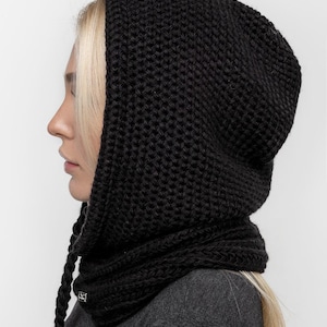 May include: A black knitted hood with a scarf-like neck warmer. The hood is made of thick, textured yarn and has long, braided ties. The neck warmer is attached to the hood. The garment is worn over a gray top.