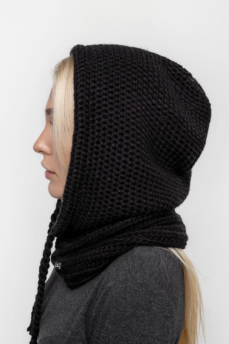 May include: A black knitted hood with a scarf-like neck warmer. The hood is made of thick, textured yarn and has long, braided ties. The neck warmer is attached to the hood. The garment is worn over a gray top.