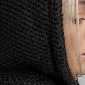 May include: Close-up of a black knitted hood. The hood is made of a thick, textured yarn, with a visible knit pattern. The hood is pulled up, partially obscuring the face of a person with blonde hair.