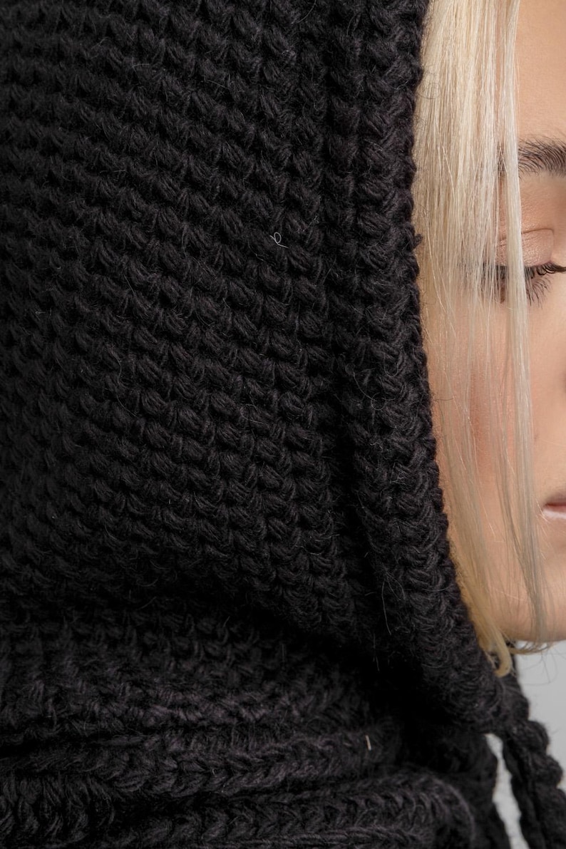 May include: Close-up of a black knitted hood. The hood is made of a thick, textured yarn, with a visible knit pattern. The hood is pulled up, partially obscuring the face of a person with blonde hair.