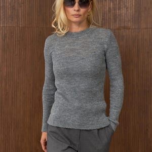 May include: A woman wearing a gray ribbed knit sweater and matching gray trousers. She is also wearing sunglasses and gold hoop earrings. The background is a brown wooden wall.
