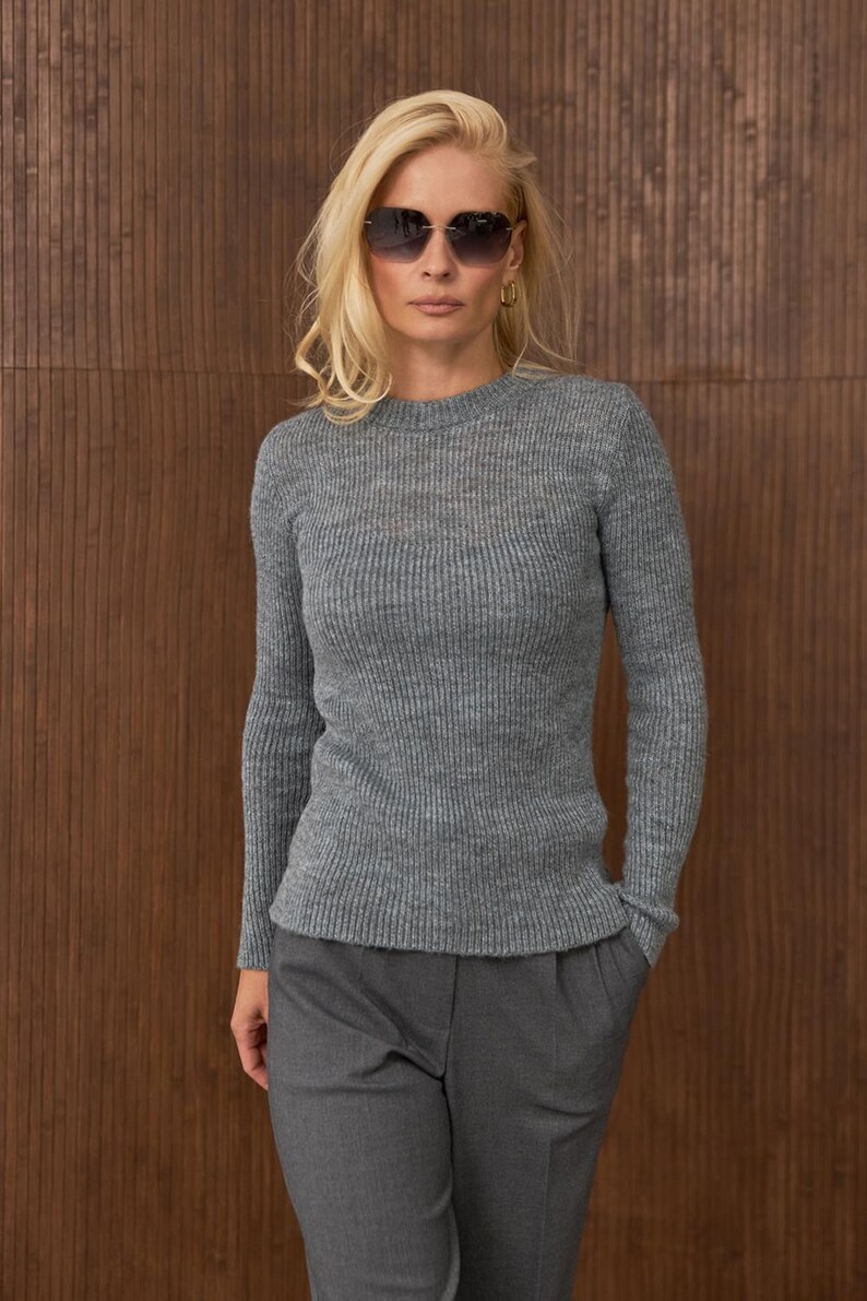 May include: A woman wearing a gray ribbed knit sweater and matching gray trousers. She is also wearing sunglasses and gold hoop earrings. The background is a brown wooden wall.
