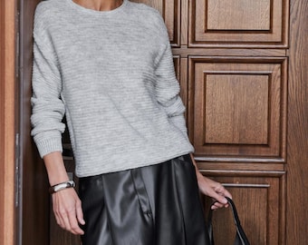 May include: A woman wearing a light gray sweater and a black leather skirt, holding a black leather tote bag. The sweater has a ribbed texture. The background is a wooden door.