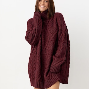 May include: A burgundy oversized cable-knit sweater dress with a turtleneck. The dress features a textured pattern and long sleeves. The model is smiling and wearing the dress. The dress is made of a thick, warm material.