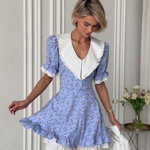 May include: A light blue floral print dress with a white collar and ruffled hem. The dress features short sleeves with white trim and a fitted bodice. The model is holding a white purse.