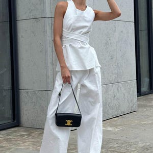 May include: A white sleeveless top with a wrap detail and wide-leg pants. The outfit is accessorized with a black shoulder bag and sunglasses.