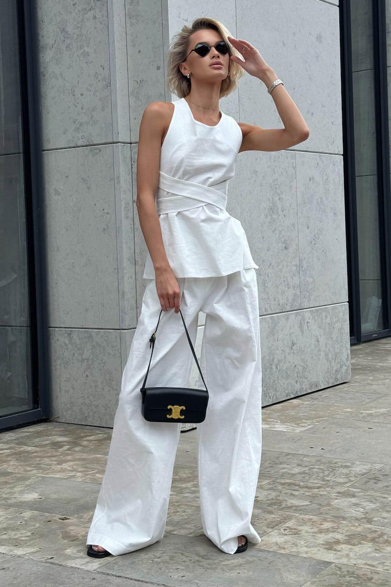 May include: A white sleeveless top with a wrap detail and wide-leg pants. The outfit is accessorized with a black shoulder bag and sunglasses.
