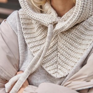 May include: A knitted hood in a light beige color. The hood is made of a thick, textured yarn and features a drawstring closure. The hood is designed to be worn over the head and neck for warmth and style.
