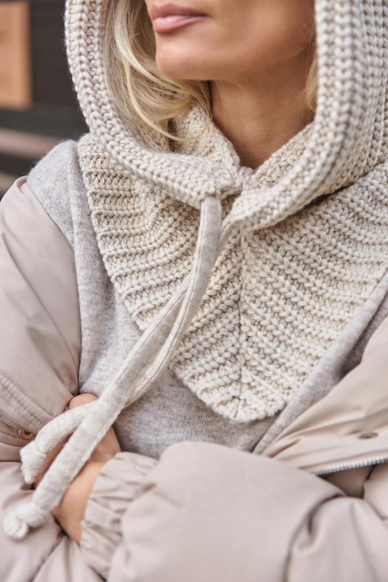 May include: A knitted hood in a light beige color. The hood is made of a thick, textured yarn and features a drawstring closure. The hood is designed to be worn over the head and neck for warmth and style.