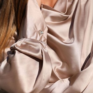 May include: A close-up of a silky, light brown blouse with a cowl neck and a single button detail on the cuff.