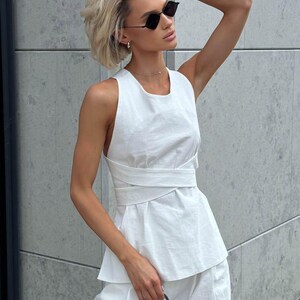 May include: A woman wearing a white sleeveless top with a criss-cross design and white pants. She is wearing black sunglasses and a silver necklace.