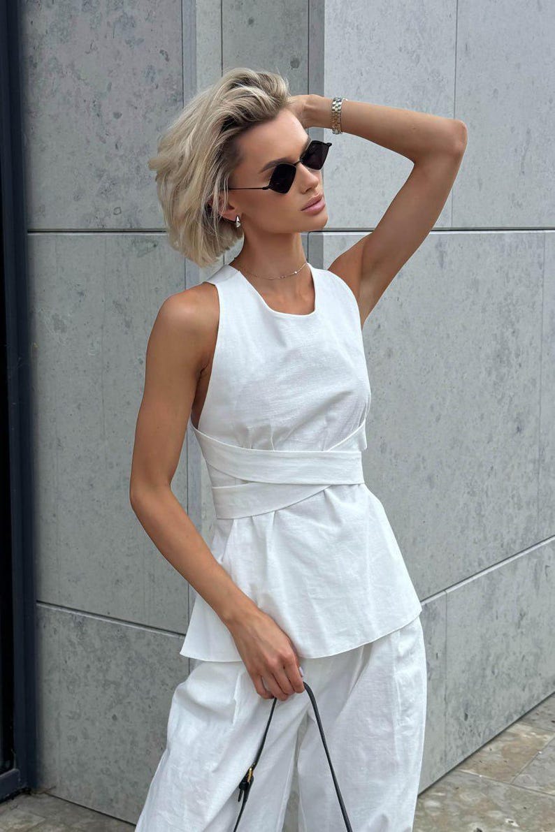 May include: A woman wearing a white sleeveless top with a criss-cross design and white pants. She is wearing black sunglasses and a silver necklace.