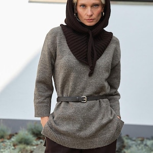 May include: A woman wearing a gray sweater, dark brown hood, and dark brown pants. The hood is knitted and tied with a drawstring. A black belt with a silver buckle cinches the waist. The background is a white wall and some greenery.