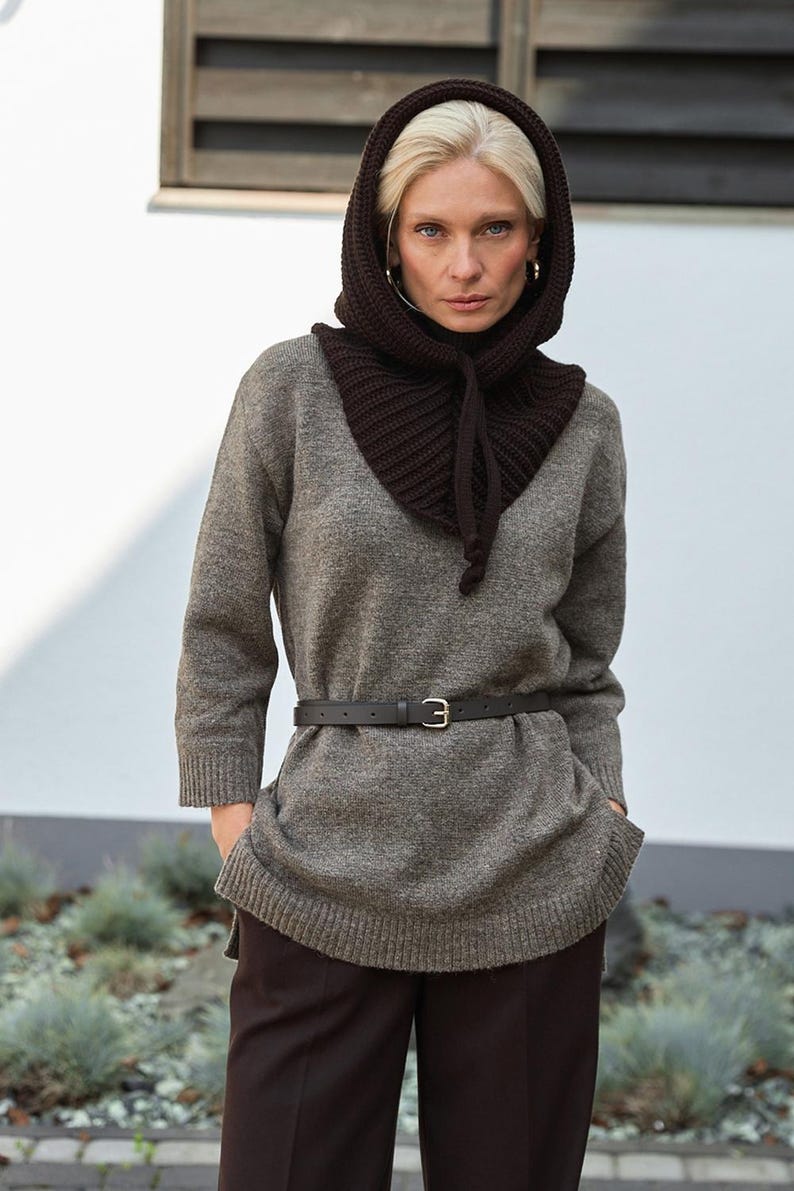 May include: A woman wearing a gray sweater, dark brown hood, and dark brown pants. The hood is knitted and tied with a drawstring. A black belt with a silver buckle cinches the waist. The background is a white wall and some greenery.