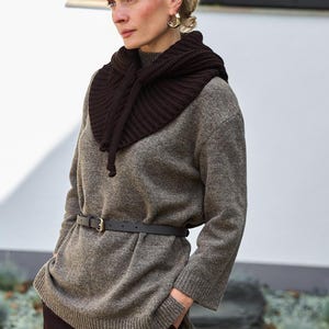 May include: A woman wearing a brown knit cowl neck warmer, a gray sweater, and a black belt. The outfit includes dark brown pants. The woman has blonde hair and gold hoop earrings.