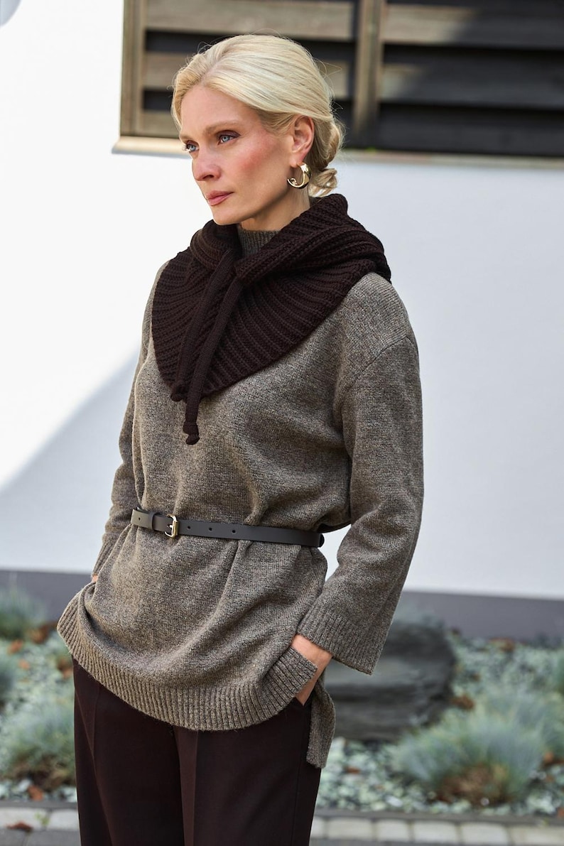 May include: A woman wearing a brown knit cowl neck warmer, a gray sweater, and a black belt. The outfit includes dark brown pants. The woman has blonde hair and gold hoop earrings.