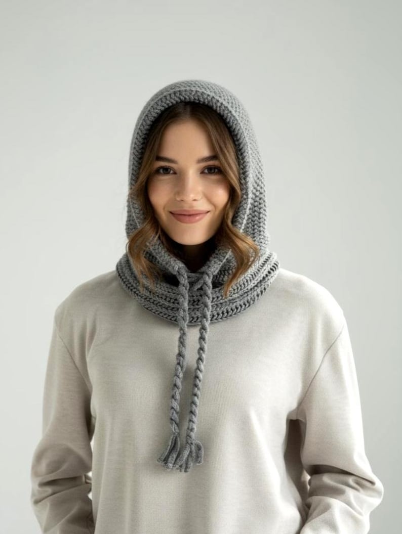 May include: A gray, knitted hood with a drawstring closure and tassels. The hood is designed to be worn around the neck and head, providing warmth and protection from the elements. The knitted texture is visible, and the hood appears to be made of a soft, comfortable material.