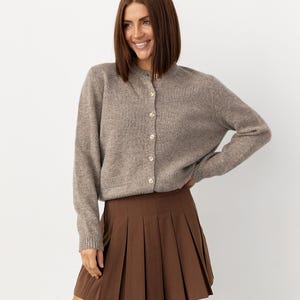 May include: A light brown cardigan with pearl buttons is paired with a brown pleated skirt. The cardigan has long sleeves and a round neckline. The skirt is short and has a classic pleated design. The outfit is simple and elegant.