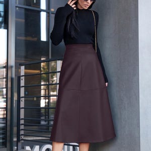 Pu&ograve; includere: Una donna indossa un maglione nero a collo alto e una gonna midi in similpelle marrone. Indossa stivaletti neri e occhiali da sole.