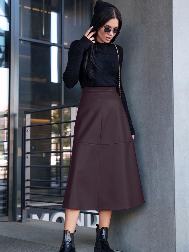 Pu&ograve; includere: Una donna indossa un maglione nero a collo alto e una gonna midi in similpelle marrone. Indossa stivaletti neri e occhiali da sole.