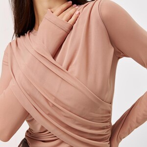 May include: A long-sleeved, blush-colored top with a draped, gathered design. The top is made of a soft, sheer fabric and has a fitted silhouette.