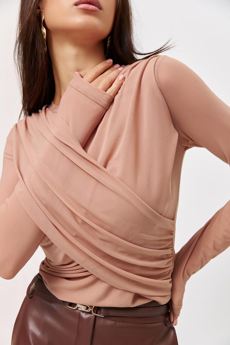 May include: A long-sleeved, blush-colored top with a draped, gathered design. The top is made of a soft, sheer fabric and has a fitted silhouette.