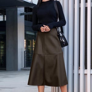 Pu&ograve; includere: Una donna indossa un maglione nero a collo alto e una gonna in similpelle marrone. Indossa un berretto da baseball nero e stivaletti neri. &Egrave; in piedi davanti a un muro bianco a listelli.