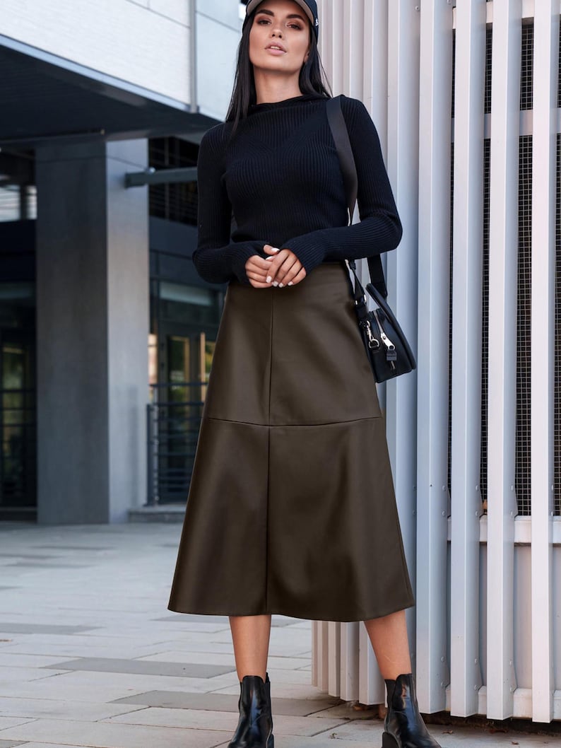 Pu&ograve; includere: Una donna indossa un maglione nero a collo alto e una gonna in similpelle marrone. Indossa un berretto da baseball nero e stivaletti neri. &Egrave; in piedi davanti a un muro bianco a listelli.