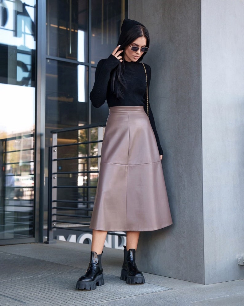 Pu&ograve; includere: Una donna indossa un maglione nero a collo alto e una gonna di pelle marrone. Indossa stivaletti neri e occhiali da sole. &Egrave; in piedi davanti a un edificio.