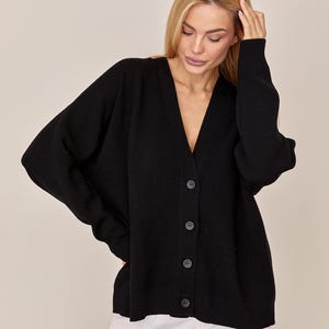 May include: A solid black long-sleeve cardigan featuring a deep V-neckline, a button-down front with four matching black buttons, ribbed cuffs, and a ribbed hem. The knit garment has a relaxed, loose fit with dropped shoulders and a smooth texture.