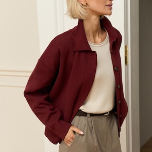 May include: A burgundy collared jacket with button closures, paired with a cream-colored top and gray trousers. A brown belt cinches the waist. The outfit is completed with gold earrings.