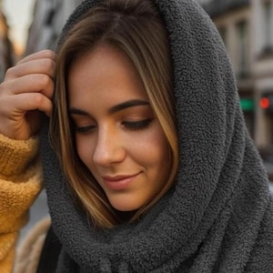 May include: A dark gray, hooded scarf made of a soft, textured material. The hood is pulled up, framing the face. The scarf appears warm and cozy, suitable for cold weather. The background is blurred, suggesting an outdoor setting.