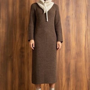 May include: A brown knit dress with a matching hood and cream-colored hood lining. The long-sleeved dress falls to mid-calf and is paired with cream-colored ankle boots. The hood has a drawstring closure.