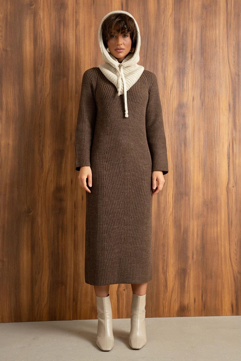 May include: A brown knit dress with a matching hood and cream-colored hood lining. The long-sleeved dress falls to mid-calf and is paired with cream-colored ankle boots. The hood has a drawstring closure.