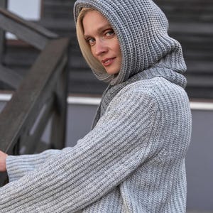 May include: A light gray knit hooded sweater with a ribbed texture. The sweater has long sleeves and a hood that covers the head and neck. The model is wearing the sweater and looking at the camera.