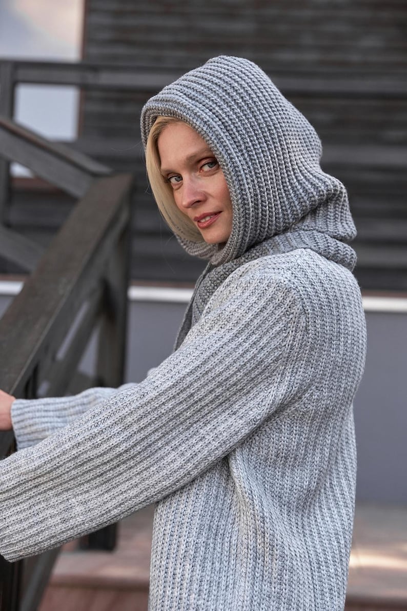 May include: A light gray knit hooded sweater with a ribbed texture. The sweater has long sleeves and a hood that covers the head and neck. The model is wearing the sweater and looking at the camera.