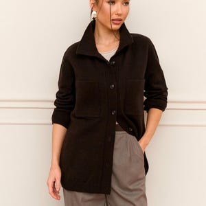 May include: A dark brown button-down jacket with two front pockets. The jacket is paired with gray trousers and a light-colored top. The model is wearing silver earrings. The outfit is suitable for a casual or business casual setting.