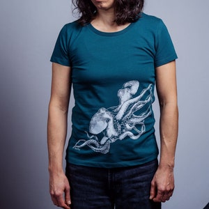 以下が含まれることがあります： 白いタコがプリントされたターコイズブルーのTシャツ。タコは8本の触手があり、丸まった状態です。Tシャツは半袖でクルーネックです。