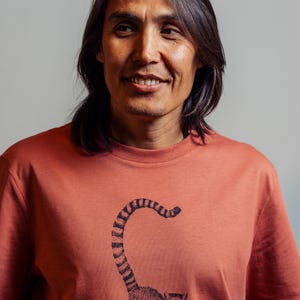 Könnte beinhalten: Ein korallenfarbenes T-Shirt mit einer detaillierten Illustration eines Lemuren mit langem, gestreiftem Schwanz. Der Lemur wird in einer stehenden Pose dargestellt. Das Shirt besteht aus weichem Material und eignet sich für Freizeitkleidung.