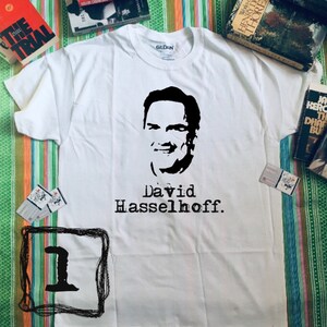 May include: White Gildan t-shirt featuring a black silhouette of David Hasselhoff's face and the name "David Hasselhoff" printed below. The shirt is surrounded by books and small cards.