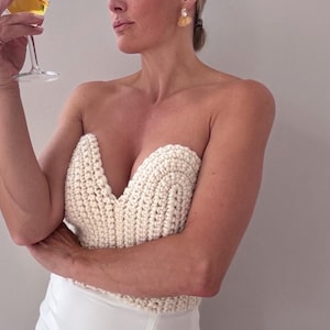 May include: A cream-colored, strapless crochet top with a deep V-neckline. The top is paired with white pants. The image also shows a person holding a martini glass with a yellow liquid and wearing gold earrings.