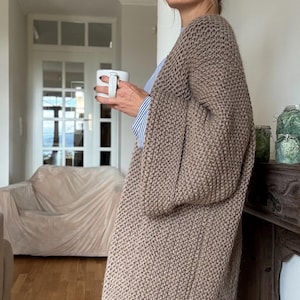 Könnte beinhalten: Langer, beigefarbener Strickcardigan mit weiten Ärmeln. Der Cardigan wird über einem blau-weiß gestreiften Hemd getragen. Eine weiße Tasse wird in der Hand gehalten. Der Hintergrund umfasst ein Sofa, eine Tür und einen Holzboden.