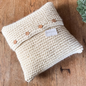 Könnte beinhalten: Ein cremefarbenes gestricktes Kissen mit einem Klappverschluss und Holzknöpfen. Das Kissen hat ein Etikett mit der Aufschrift "Stella Noctium - Berlin".