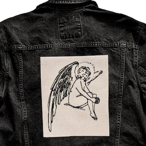 Peut inclure: Veste en jean noire avec un patch rectangulaire au dos. Le patch présente une illustration en noir et blanc d'un ange avec des ailes. La veste a un design classique avec des fermetures à boutons, des poches et un col.