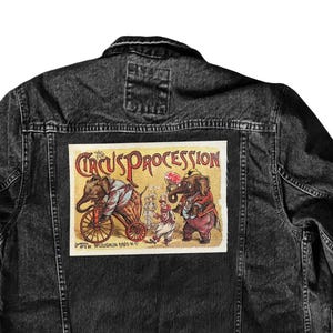 May include: A black denim jacket with a vintage circus procession patch on the back. The patch features a colorful illustration of elephants, a wagon, and people.