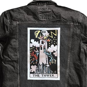 May include: A black denim jacket with a tarot card graphic on the back. The graphic features a tower being struck by lightning, with the words "THE TOWER" at the bottom. The jacket has a button-up front and two chest pockets.