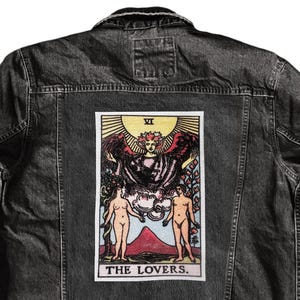 May include: Black denim jacket with a rectangular patch on the back featuring a tarot card illustration. The card depicts two figures and the text "THE LOVERS."