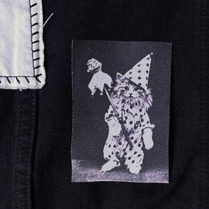 May include: Black denim jacket with a whimsical patch featuring a cat dressed as a jester, holding a staff. The patch is black and white with a polka dot pattern. The jacket has two black buttons.