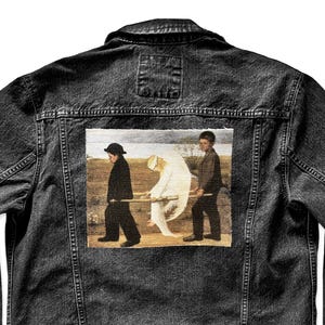 May include: Black denim jacket with a button-down front, chest pockets, and a classic collar. The back features a rectangular patch with a painting of three figures carrying a white form across a landscape.