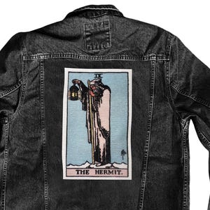 May include: Dark grey denim jacket with a tarot card design on the back. The patch shows a robed figure with a lantern on a light blue backdrop. The text "THE HERMIT" is printed at the bottom of the patch, below the illustration.